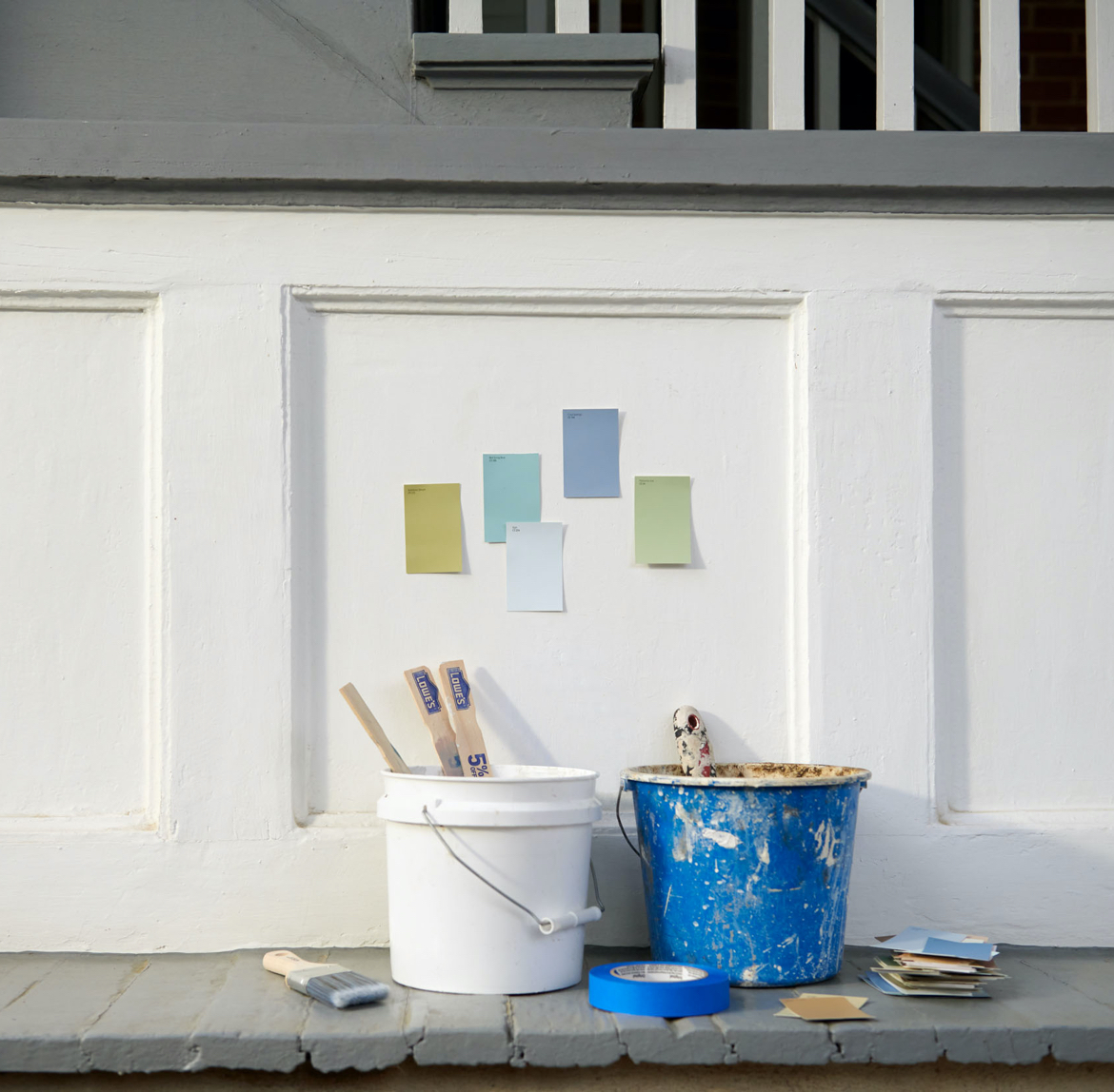 Pared de paneles blanca debajo de escaleras con muestras de color. Cubetas de pintura desordenadas con agitadores de pintura y muestras debajo.