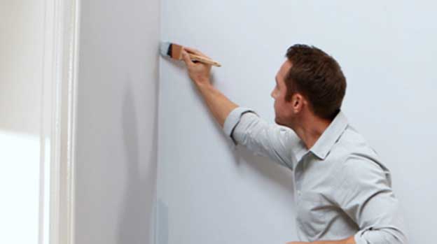 Man in button-down shirt holds brush with white paint and is painting the edge of the wall at the corner.