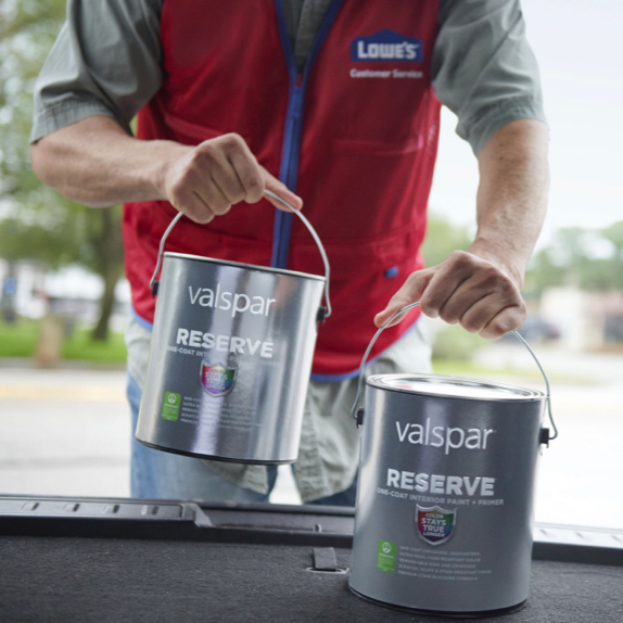 Representante de servicio al cliente de Lowe's® con chaleco rojo cargando dos latas de pintura Valspar® Reserve® en la cajuela de un auto.