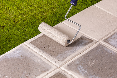 Concrete walkway across grass lawn being sealed by roller with white sealer.
