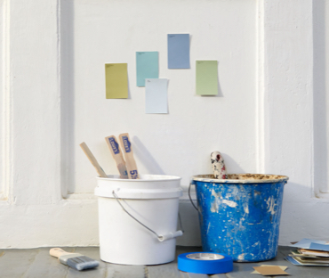 Cubetas de pintura blanca y azul, brocha de pintura, cinta azul y muestras en el piso. Muestras en pared blanca. Consulta de color