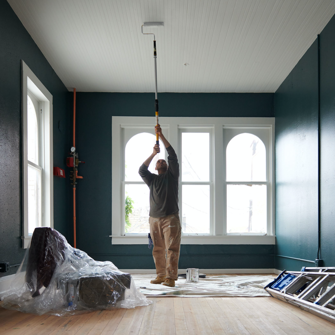 Persona en habitación grande con ventanas que usa un rodillo con una vara de extensión para pintar el techo.