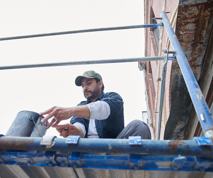 Persona en andamio en frente de un edificio de ladrillo que sumerge una brocha en una lata.
