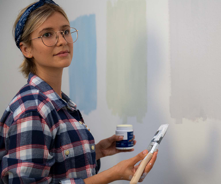 DIY painter wearing flannel and oversized round metal glasses holds paint brush. Wall has three paint samples.