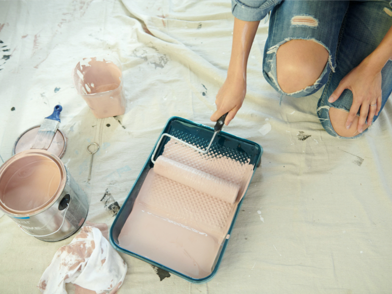 Person holding  roller in tray on top of drop cloth  with a can, brush and rag in foreground.