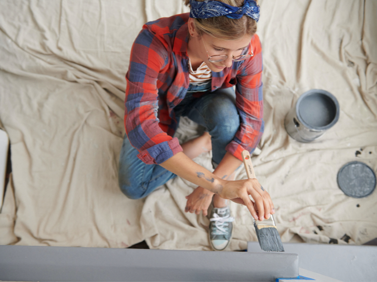 Persona sentada en un paño, aplicando color gris a una pared con una lata abierta llena de pintura junto a la persona.