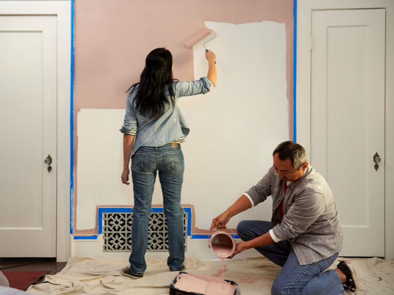 Persona usando un rodillo para aplicar color a pared entre dos puertas con otra persona que vierte una lata a una bandeja.